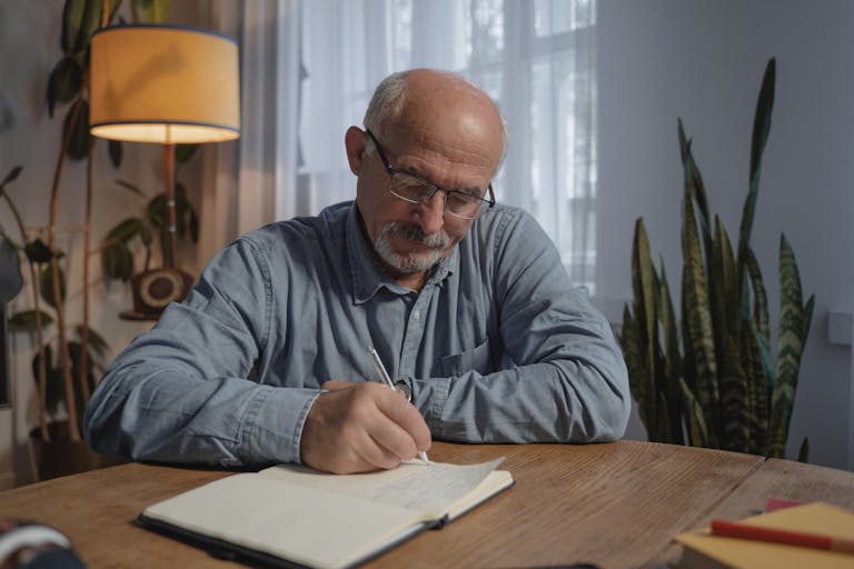 A senior adult man writing in a notebook at a desk in a cozy home office setting.