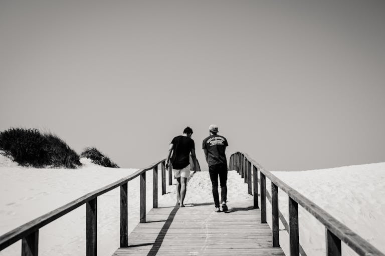 Two men walk on a wooden boardwalk in a snowy beach setting, creating a serene grayscale scene.